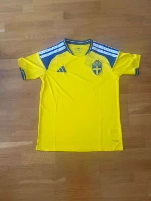 Svensk landslagströja Adidas gul - Gul svensk landslagströja från Adidas med blå detaljer och tre vita ränder på axlarna. Broderad svensk landslagssköld på bröstet och Adidas-logga. Tillverkad i lätt och ventilerande material, perfekt för fotboll eller träning.