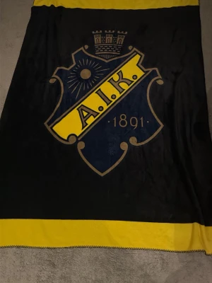 AIK filt svart och gul - Mjuk filt i svart och gult med stort AIK-emblem och årtalet 1891 tryckt på framsidan. Perfekt för dig som vill visa ditt AIK-supporterhjärta hemma eller på matchen. Tillverkad i skönt material och har tydliga klubbfärger.