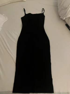 Svart långklänning med smala axelband - Stilren svart långklänning med smala axelband och figurnära passform. Klänningen har en enkel och elegant design som framhäver siluetten. Perfekt för dig som gillar minimalistisk stil och vill ha en klassisk look.