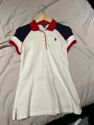 Vit pikéklänning från U.S. Polo Assn. - Vit pikétröja från U.S. Polo Assn. med röda och marinblå detaljer. Tröjan har korta ärmar, röd krage och knappslå framtill. Liten broderad logga på bröstet och smal passform. Tillverkad i bomull med lite stretch för extra komfort. Köpt i USA.