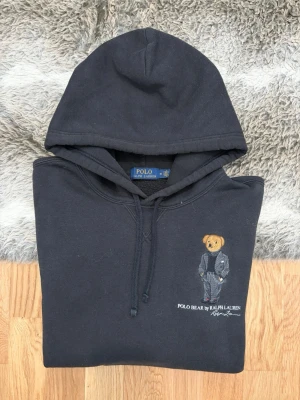 Svart Polo Ralph Lauren Bear Hoodie - Storlek M - Hej! 👋 Säljer en riktigt snygg och ikonisk svart Polo Ralph Lauren "Polo Bear"-hoodie i storlek M. 🐻✨ Ett måste för varje samling! Tröjan är i mycket gott skick och välvårdad. Det kan förekomma något enstaka litet nopprigt parti (helt naturligt för materialet), men det är inget som stör helhetsintrycket. 100% äkta med tvättlappar kvar (ingen QR-kod på denna modell). En riktig stilikon som passar till allt. Fraktar snabbt! 🚛📦💬💯 