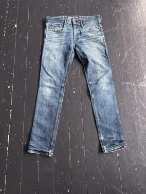 Blå Replay jeans - Säljer ett par blå jeans från Replay med snygga slitningar och klassisk femficksdesign. Jeansen har raka ben och detaljer som Replay-logga på fickan. Perfekta för en avslappnad och trendig stil.