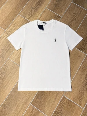 Vit t-shirt från Saint Laurent - Vit t-shirt från Saint Laurent med klassisk rund hals och svart YSL-logga broderad på bröstet. Tillverkad i mjuk bomull och har en stilren, normal passform. Perfekt för en clean och trendig look.