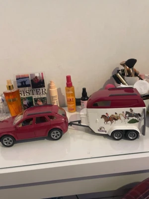 Leksaksbil med hästtransport - Säljer en röd leksaksbil i plast med beige inredning och öppningsbar baklucka. Medföljer ett vitt och rött hästsläp dekorerat med hästmotiv på sidorna. Perfekt för dig som gillar hästar och bilar! Har lite klister kvar från gammla klistermärken men går säkert att skrapa bort.