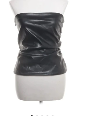 Svart axelbandslös tubtopp i skinnimitation - Snygg svart axelbandslös tubtopp i skinnimitation. Toppen har en slät och glansig yta med en tight passform som ger en edgy look. Perfekt att styla med jeans eller kjol för en trendig outfit.