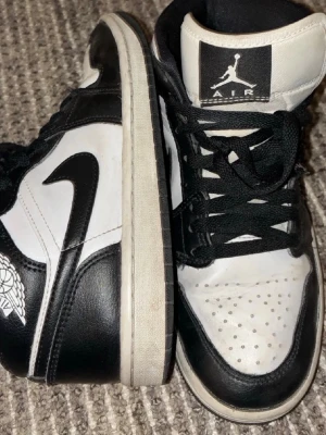 Svarta och vita Air Jordan 1 High - Säljer ett par klassiska Air Jordan 1 High i svart och vitt läder. Skorna har den ikoniska swoosh-loggan på sidan, perforerad tåbox och högt skaft. Perfekta för dig som gillar streetwear och vill sticka ut med en tidlös sneaker.