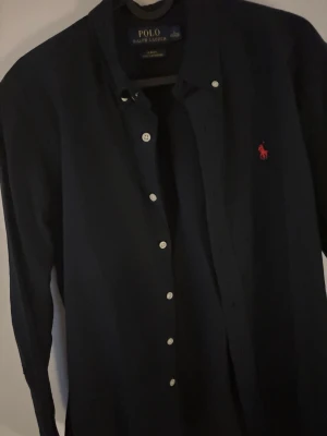 Mörkblå skjorta från Polo Ralph Lauren - Klassisk mörkblå skjorta från Polo Ralph Lauren med vit knappstängning och ikonisk röd logga broderad på bröstet. Skjortan har lång ärm och tidlös design, perfekt för en stilren look. Sitter litet rekommenderas på folk som e 170-175