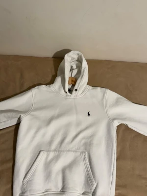 Vit hoodie från Polo Ralph Lauren - Säljer en klassisk vit hoodie från Polo Ralph Lauren med broderad logga på bröstet. Hoodien har en stor huva med metallöglor, ribbade muddar och en praktisk magficka. Perfekt för dig som gillar stilren och tidlös design.
