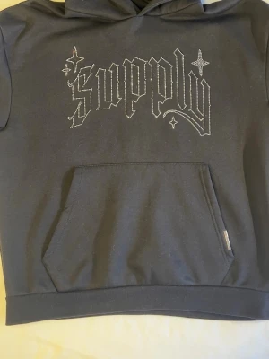 Svart hoodie med strasstext Supply - Svart hoodie med huva och magficka. På bröstet står det 'supply' i gotisk stil med strasstenar, och ärmen har flera stjärnor i strass. Perfekt för dig som gillar streetwear och detaljer som sticker ut.