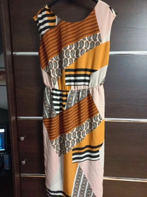 Mönstrad långklänning i orange och beige - Unik klänning med coolt grafiskt mönster i orange, beige, svart och vitt. Klänningen har breda axelband, rund halsringning fram och v-ringad rygg. Midjan är markerad med resår och tyget är mjukt och skönt, perfekt för varma dagar.