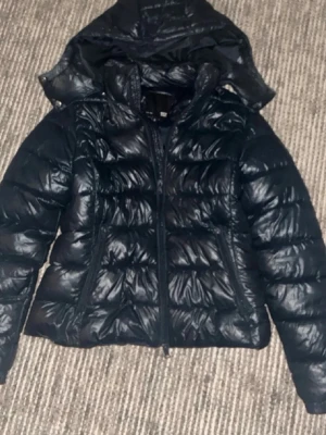 Svart pufferjacka med huva - Snygg svart pufferjacka med glansig finish och huva. Jackan har dragkedja framtill och två sidofickor. Perfekt för kalla dagar och ger en modern look. Fylld med vaddering för extra värme och komfort.