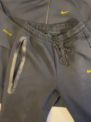 Svart NOCTA hoodie från Nike - Svart hoodie från Nike NOCTA-kollektionen med gul logga på bröstet och NOCTA-tryck på ärmen. Hoodien har dragkedja framtill, två fickor med detaljer och en stor huva. Materialet är mjukt och bekvämt, perfekt för en streetwear-look.