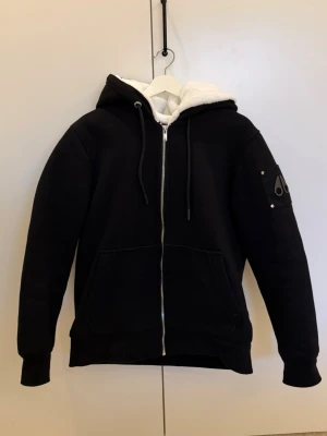 Svart hoodie Moose Knuckles med foder - Snygg svart hoodie från Moose Knuckles med dragkedja, vit fluffig foder på insidan och huva med snören. Har fickor framtill och en logga på ärmen. Perfekt för dig som vill ha en varm och stilren hoodie med exklusiv känsla.