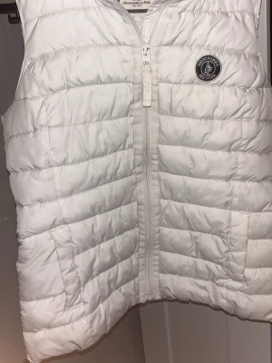Vit dunväst från Abercrombie & Fitch - Snygg vit dunväst från Abercrombie & Fitch med quiltad design och dragkedja framtill. Västen har två fickor och en rund logotyp-patch på bröstet. Perfekt att slänga över en hoodie eller tröja för extra värme och stil.