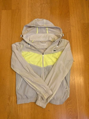 Beige och gul vindjacka från Lacoste - Snygg beige vindjacka från Lacoste med gul panel över bröstet och huva. Jackan har dragkedja framtill, två sidofickor med dragkedja och elastiska muddar vid ärmslut och nederkant. Tillverkad i lätt syntetmaterial, perfekt för blåsiga dagar.