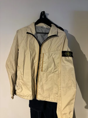 Beige Stone Island vindjacka - Beige vindjacka från Stone Island med ikonisk svart logotyp-patch på vänster ärm. Jackan har dragkedja framtill, hög krage och är tillverkad i ett lätt, prassligt material. Enkel design med diskreta sömmar och en bröstficka med dragkedja. Perfekt för dig som gillar streetwear.