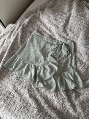 Ljusgrön omlottkjol med volang - Supersöt ljusgrön kortkjol med ”blommigt” mönster, omlottmodell och volangkant. Kjolen har en knytning i midjan och inbyggda shorts under för extra komfort. Använd någon enstaka gång men har tyvärr fått en fläck (se sista bilden), men inget som märks av!