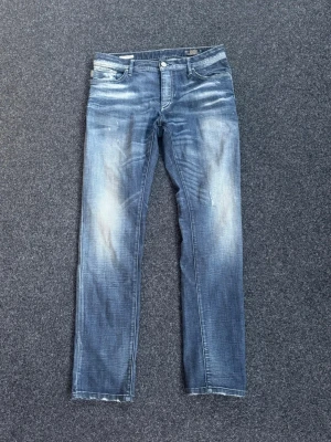 Blå slim fit jeans från Jack and Jones  - Snygga blå slim fit jeans från Jack and Jones med slitna detaljer och ljusa partier framtill. Klassisk femficksmodell med läderpatch bak i midjan. Jeansen har en modern tvätt och raka ben, perfekta för en avslappnad streetstil.                              Storlek 32-32, dock har storleks lappen försvunnit.