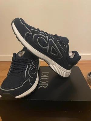 Marinblå CD  sneakers med vita detaljer - Snygga marinblå CD sneakers med vita och grå detaljer. Skorna har en sportig design med mesh och mockaliknande material, samt en tjock vit sula. Stora CD -loggor på sidan och klassisk snörning framtill. Perfekta för dig som vill ha något exklusivt och trendigt.