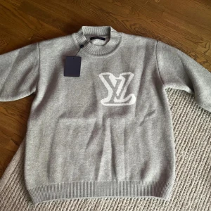 Grå stickad tröja Louis Vuitton - Snygg grå stickad tröja från Louis Vuitton med stort vitt LV-logo framtill. Tröjan har ribbad krage, ärmslut och nederkant. Perfekt för dig som vill ha en exklusiv och stilren look med lyxig känsla.