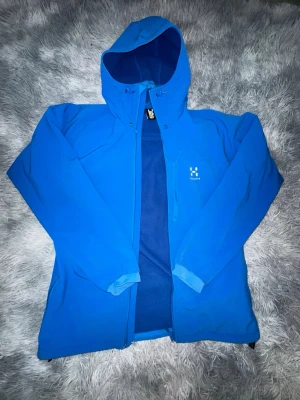 Blå sportjacka med huva från Haglöfs - Blå sportjacka från Haglöfs med justerbar huva och dragkedja framtill. Jackan har en bröstficka med dragkedja och är gjord i ett slitstarkt, vindtätt material som passar perfekt för träning och utomhusaktiviteter.