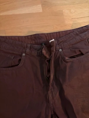 Bruna jeans med raka ben - Säljer ett par bruna jeans med klassisk femficksdesign och raka ben. Jeansen har dragkedja och knapp framtill samt bakfickor. Materialet är bomull och färgen är mörkbrun, perfekt för dig som vill ha en stilren och enkel look.