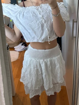 Vit offshoulder croppad blus - Söt vit offshoulder blus med broderade detaljer och croppad passform. Blusen har resår vid axlarna och midjan samt dekorativ knytning framtill. Perfekt för sommarens alla tillfällen och ger en romantisk vibe.