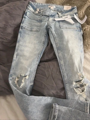 Ljusblåa bootcut jeans med slitningar - Säljer ett par ljusblåa bootcut jeans med slitna detaljer på båda knäna och råa kanter. Jeansen har låg midja och stora fickor framtill. Perfekta för en avslappnad och trendig look. Skriv för mer bilder, pris går att diskuteras🫶🏻