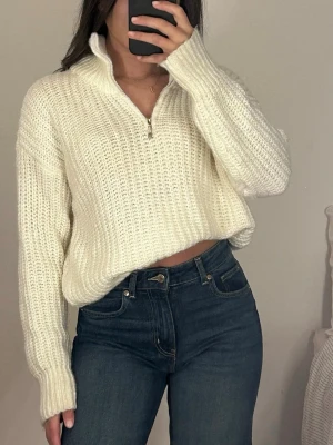 Vit stickad halvzip tröja - Mysig vit stickad tröja med halvzip och hög krage. Tröjan har en grovstickad struktur, långa ärmar och ribbade muddar. Perfekt för lager-på-lager och chill dagar. Materialet ser ut att vara mjukt och värmande. M