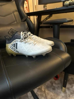 Adidas X 16.1 AG - Snygga fotbollsskor från Adidas i vitt med svart logga och guldfärgad häl. Skorna har låg profil, snörning och mönstrad ovandel för extra grepp på planen. Perfekta för dig som vill sticka ut på matchen och få bra bollkontroll.