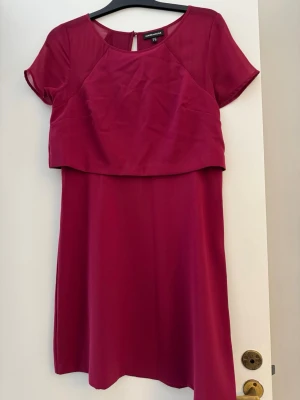 Mörkrosa kortklänning från Warehouse - Snygg mörkrosa kortklänning från Warehouse med korta ärmar och rund halsringning. Klänningen har en modern lager-på-lager-look upptill och är tillverkad i ett lätt, slätt material som ger ett stilrent intryck.