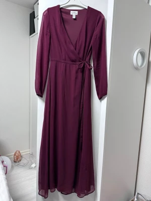 Vinröd omlottklänning från NLY  - Vinröd långklänning från NLY One med omlottmodell och knyt i midjan. Klänningen har långa ärmar och v-ringad halsringning. Tillverkad i ett lätt, flowy material som ger en elegant siluett. Storlek 36