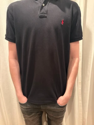 Svart pikétröja från Ralph Lauren - Klassisk svart pikétröja från Ralph Lauren med korta ärmar och röd broderad logga på bröstet. Tröjan har krage med knappar och är gjord i mjuk bomull som känns skön mot huden. Perfekt till jeans eller shorts för en clean look. Storlek L, modellen är 185 cm och jag skulle säga att den passar någon som är mellan 185-195 men det beror på vikt.