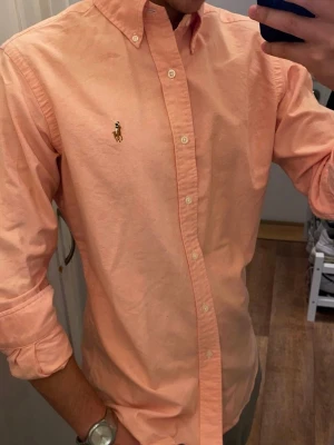 Rosa Skjorta från Ralph Lauren Oxford (Slim Fit) - Klassisk persikofärgad skjorta från Polo Ralph Lauren med button-down krage och broderad logga på bröstet. Skjortan har långa ärmar, knappar framtill och är tillverkad i bomull. Perfekt för en stilren och avslappnad look. Storlek Small Slim fit. Snabb Leverans!