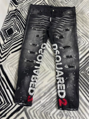 Svarta Dsquared2 jeans med tryck - Säljer ett par svarta jeans från Dsquared2 med slitningar, vita färgstänk och stort vitt Dsquared2-syd längs benen. Jeansen har fem fickor, knappgylf och röda detaljer i trycket. Snygg streetstil och trendig passform. Pris kan diskuteras 