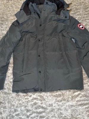 Mörkgrå dunjacka från Canada Goose - Snygg mörkgrå dunjacka från Canada Goose med klassisk patch på ärmen. Jackan har hög krage, huva och dragkedja med knappslå. Två stora fickor framtill och en stilren, rak passform. Perfekt för kalla vinterdagar.