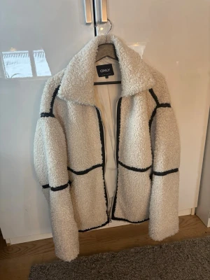 Vit teddyjacka från ONLY - Säljer en fluffig vit teddyjacka från ONLY med svarta kontrastsömmar. Jackan har bred krage och oversized passform. Ny pris 899
