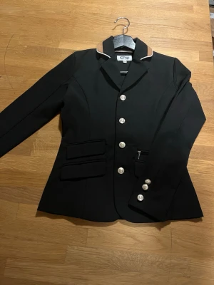 Svart ridkavaj från CRW - Snygg svart ridkavaj från CRW med knappar framtill och på ryggen. Kavajen har en beige och vit detalj på kragen, figursydd passform och flera fickor framtill. Perfekt för dig som vill ha en stilren look till ridningen. Barn/Junior