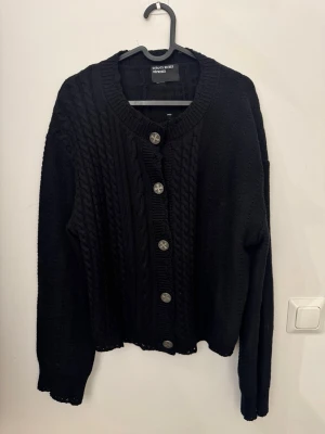 Svart kabelstickad kofta/cardigan Enfants Riches Deprimes - Kabelstickad svart kofta från Enfants Riches Deprimes med grova flätmönster och fem dekorativa metallknappar framtill. Koftan har rund halsringning, ribbade muddar och en lite boxig passform med kroppad midja. Perfekt för lager på lager och en stilren look.