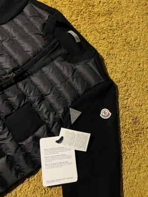 Svart cardiganjacka från Moncler - Snygg svart cardiganjacka från Moncler med quiltad, vadderad front och stickade ärmar. Jackan har två stora fickor framtill och dragkedja. Moncler-logga på ärmen och ribbade muddar vid ärmslut och nederkant. Perfekt för lager på lager och streetstyle. Allt medföljer inklusive dustbag,kvitto,etiketter