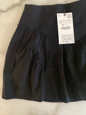 Svart volangkjol från Gina Tricot - Kort svart kjol från Gina Tricot med bred linning och volangdetaljer. Kjolen har en luftig passform och är tillverkad i ett mjukt bomullstyg som känns skönt mot huden. Perfekt att styla med en enkel topp eller hoodie. Shorts under. 