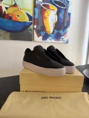 Svarta Axel Arigato sneakers - Snygga svarta sneakers från Axel Arigato med minimalistisk design och vit sula. Skorna är i slätt läder med diskret logga i guld på sidan och elastiska paneler för enkel påtagning. Perfekta för dig som gillar stilrena och moderna skor.