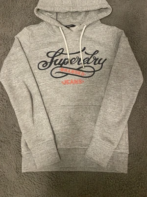 Grå hoodie från Superdry - Snygg grå hoodie från Superdry med stor logga i svart och rött framtill. Tröjan är i väldigt bra skick och har inga defekter, fortfarande mjuk på insidan! Storlek XS men skulle säga att den passar på någon som är runt 160. Hör gärna av dig vid frågor eller funderingar!😁