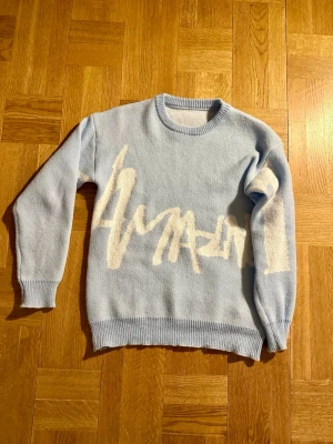 Ljusblå tröja - Manfinity - Säljer en ljusblå stickad tröja med vit grafisk text över bröstet. Tröjan har rund halsringning, ribbade muddar och nederkant. Perfekt för dig som gillar en avslappnad och trendig stil.