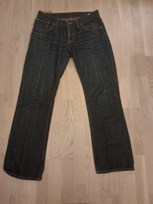 Mörkblå jeans från Nudie Jeans - Säljer ett par mörkblå jeans från Nudie Jeans med klassisk femficksmodell och raka ben. Jeansen har kontrastsömmar och Nudie-logga på bakfickan. Tillverkade i slitstark denim, perfekta för en avslappnad och stilren look.
