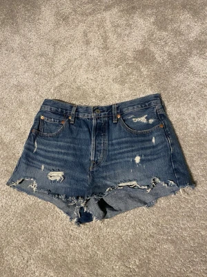 Levi's blå jeansshorts med slitningar 501’s - Säljer ett par klassiska blå jeansshorts från Levi's med rå kant och slitna detaljer. Shortsen har fem fickor, knappgylf och den ikoniska Levi's-lappen bak. Perfekta för sommaren och ger en avslappnad look. Det är 501’s