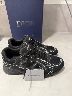 Svarta Dior b30  - Snygga svarta Dior sneakers med vita kontrastdetaljer och tydlig logga på sidan. Skorna har en sportig siluett, svarta skosnören och mesh- samt mockaliknande material. Perfekta för dig som vill ha en exklusiv och trendig look.