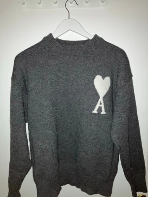 Grå stickad tröja från Ami Paris - Säljer en grå stickad tröja från Ami Paris med deras ikoniska vita hjärta och A på bröstet. Tröjan har rund halsringning, ribbade muddar och är gjord i mjukt material som känns skönt mot huden. Perfekt för dig som gillar stilrena och trendiga plagg.