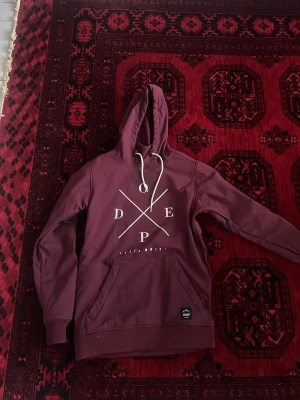 skidjacka från Dope - Säljer en vinröd skidjacka  med huva från Dope. Jackan har vit tryckt logga på bröstet, snörning i huvan och en stor magficka. Materialet är mjukt och jackan har ribbade muddar vid ärmslut och nederkant. Perfekt för en chill look.