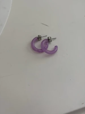 Lila hoops i plast - Säljer ett par snygga örhängen i form av små hoops i genomskinlig lila plast. De har metallstift och en enkel, modern design som passar till många olika stilar. Perfekta för dig som gillar färgglada accessoarer! Aldrig använt från Kappahl.